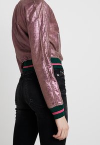 Metaliczna różowa kurtka o kroju cropped z prążkowanym zielonym i różowym pasem, zapinana na zamek, z długimi rękawami, połączona z czarnymi dżinsami.