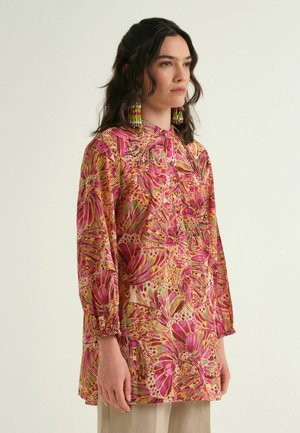 Frau trägt eine langärmelige pink-orange Schmetterlingsmuster-Bluse mit Quastenohrringen und steht vor einem einfarbigen hellen Hintergrund.