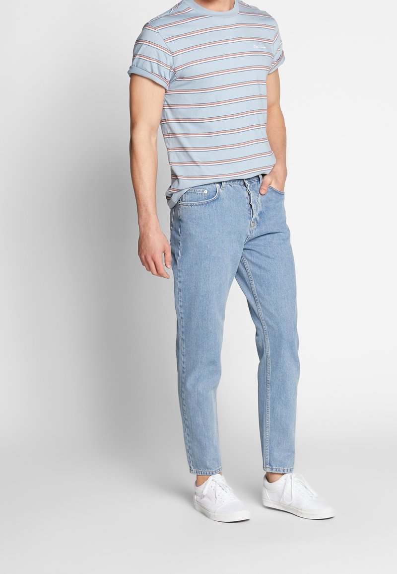 Homme portant un t-shirt rayé bleu clair, un jean bleu clair et des baskets blanches, debout avec une main dans la poche.