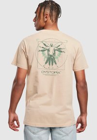 Beige cotton t-shirt featuring a green graphic design on the back, depicting a figure with wings inside a geometric pattern.