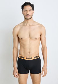 Schwarze Boxershorts mit einem breiten orangen Bund, der das Puma-Logo trägt; der Stoff wirkt glatt und hat einen figurschmeichelnden Schnitt. Das Modell steht ohne Shirt.