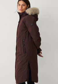 Bruine parka met een capuchon met bontkraag, lange mouwen, zakken met rits en een ritssluiting aan de voorkant. Gladde stof met een matte afwerking.