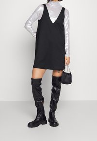 Schwarzes ärmelloses Kleid mit tiefem V-Ausschnitt, kombiniert mit einem schimmernden silbernen Langarmoberteil, hochgeschnittenen schwarzen Lederstiefeln und einer kleinen Handtasche.