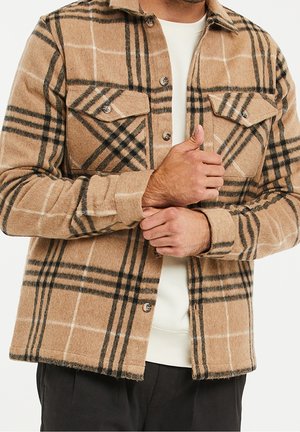 Homme portant une veste à carreaux beige et noire à boutons sur une chemise blanche, ajustant le poignet gauche avec la main droite.