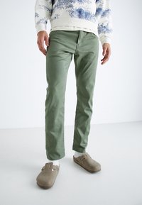 Mies, joka on pukeutunut vaaleanvihreisiin housuihin, valkoisiin sukkiin, beigeihin liukuviin kenkiin ja valkoiseen huppariin, jossa on sinisiä maisemakuviota valkoisella taustalla.