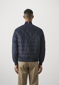 Man i en marinblå vadderad jacka med diamant- och horisontell sömnad, stående med händerna nere, i beige byxor och vänd bort från kameran.