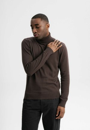 Brauner Rollkragenpullover mit gerippten Bündchen und Saum, figurbetontes Design, getragen von einer Person mit kurzen Haaren, schwarzen Hosen und dezentem Handplatz.