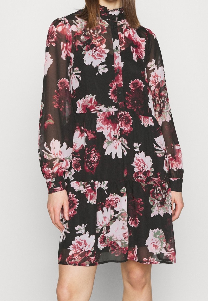Robe noire à fleurs en tissu léger et transparent avec des motifs de fleurs roses et blanches. Elle comporte un col rond et des manches longues.