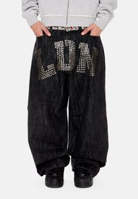 Άτομο που φορά χαλαρό μαύρο τζιν παντελόνι με μεγάλα ασημένια τρουκς που σχηματίζουν τη λέξη "LDRK" πάνω στις εμπρόσθιες τσέπες και μαύρα αθλητικά παπούτσια.