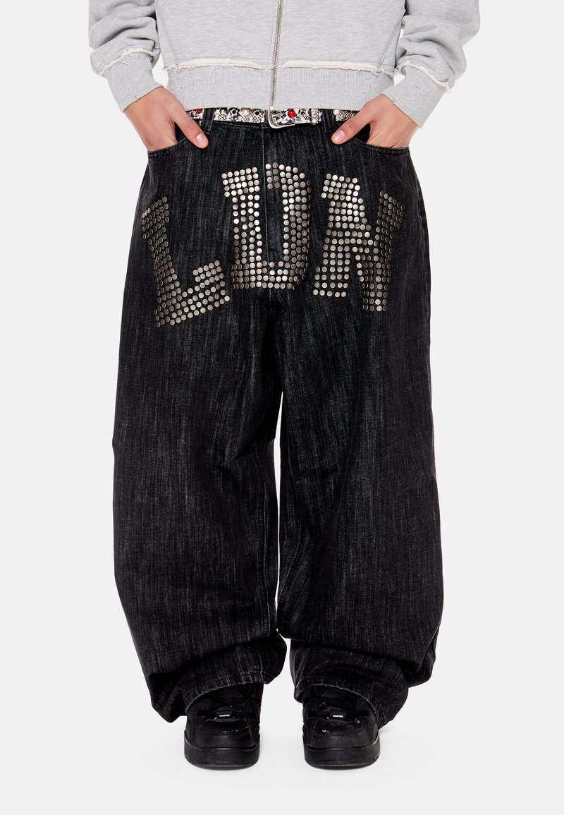 Άτομο που φορά χαλαρό μαύρο τζιν παντελόνι με μεγάλα ασημένια τρουκς που σχηματίζουν τη λέξη "LDRK" πάνω στις εμπρόσθιες τσέπες και μαύρα αθλητικά παπούτσια.