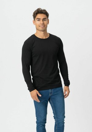 Junger Mann steht lächelnd da, trägt ein schwarzes Langarmshirt und blaue Jeans vor einem schlichten hellen Hintergrund.