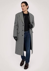 Grå ullkappa med skräddarsydd passform, stora slag och knappstängning, kombinerad med svart skjorta, blå jeans och mörkbruna skor.