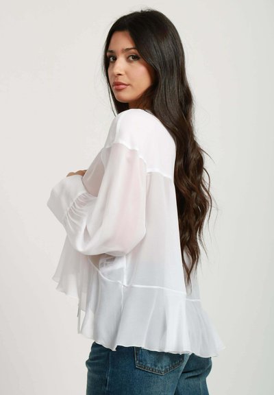 Blusa bianca in chiffon con maniche lunghe, orlo arricciato e vestibilità rilassata. Abbinata a jeans in denim blu, modello di profilo a destra.