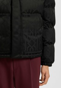 Giubbotto a vento nero con un sottile motivo geometrico, tasche piatte e polsini elasticizzati. Abbinato a pantaloni bordeaux.