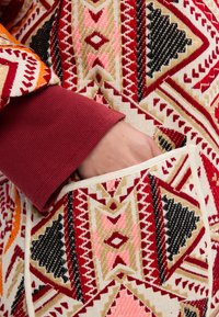 Textúrált szövet geometriai mintával vibráló piros, narancs, rózsaszín és fekete színekben. Látható kézipocket krém szegéllyel, piros ujjszárral.