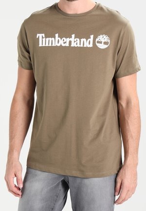 Mand iført en olivengrøn Timberland T-shirt med hvidt logo, kombineret med lysegrå jeans, står mod en neutral baggrund.