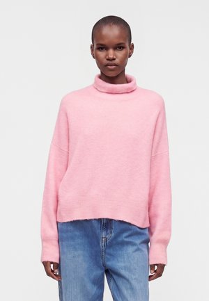 Personne portant un pull à col roulé rose pâle et un jean bleu, debout devant un fond blanc uni.