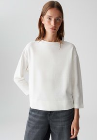 Giovane donna con capelli castano chiaro che indossa un top bianco a maniche lunghe con texture e jeans grigio scuro, in piedi contro uno sfondo chiaro e semplice.