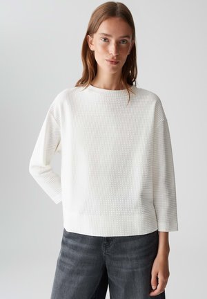 Jonge vrouw met lichtbruin haar, gekleed in een getextureerde witte longsleeve en donkergrijze jeans, staand tegen een lichte effen achtergrond.