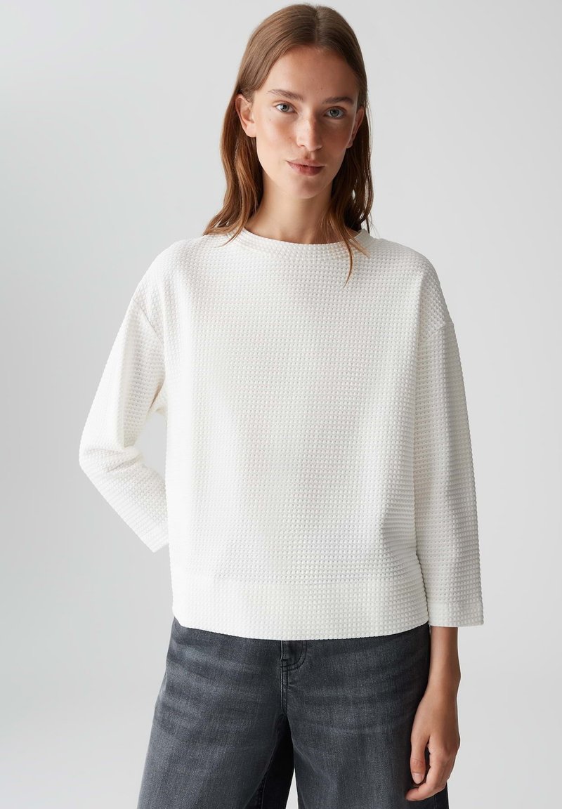 Giovane donna con capelli castano chiaro che indossa un top bianco a maniche lunghe con texture e jeans grigio scuro, in piedi contro uno sfondo chiaro e semplice.