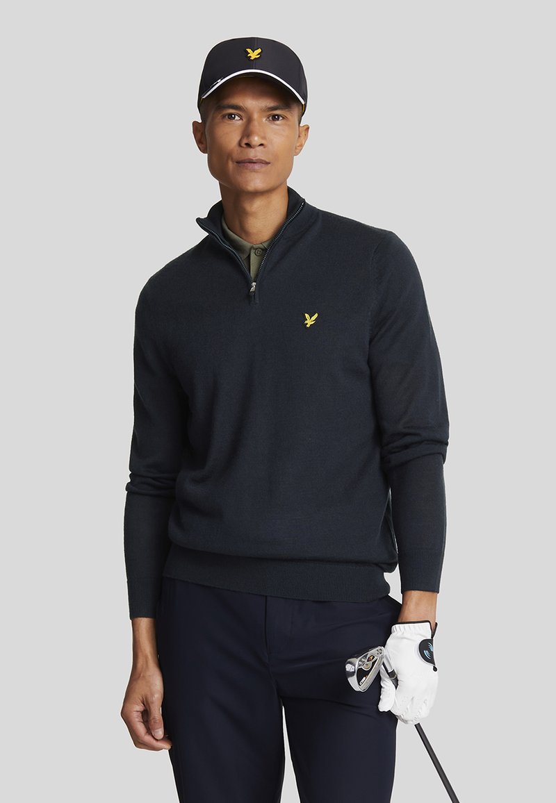 Navy pullover sweatshirt with a quarter-zip neckline, featuring a small yellow logo. Model wears a black cap and holds a golf club.