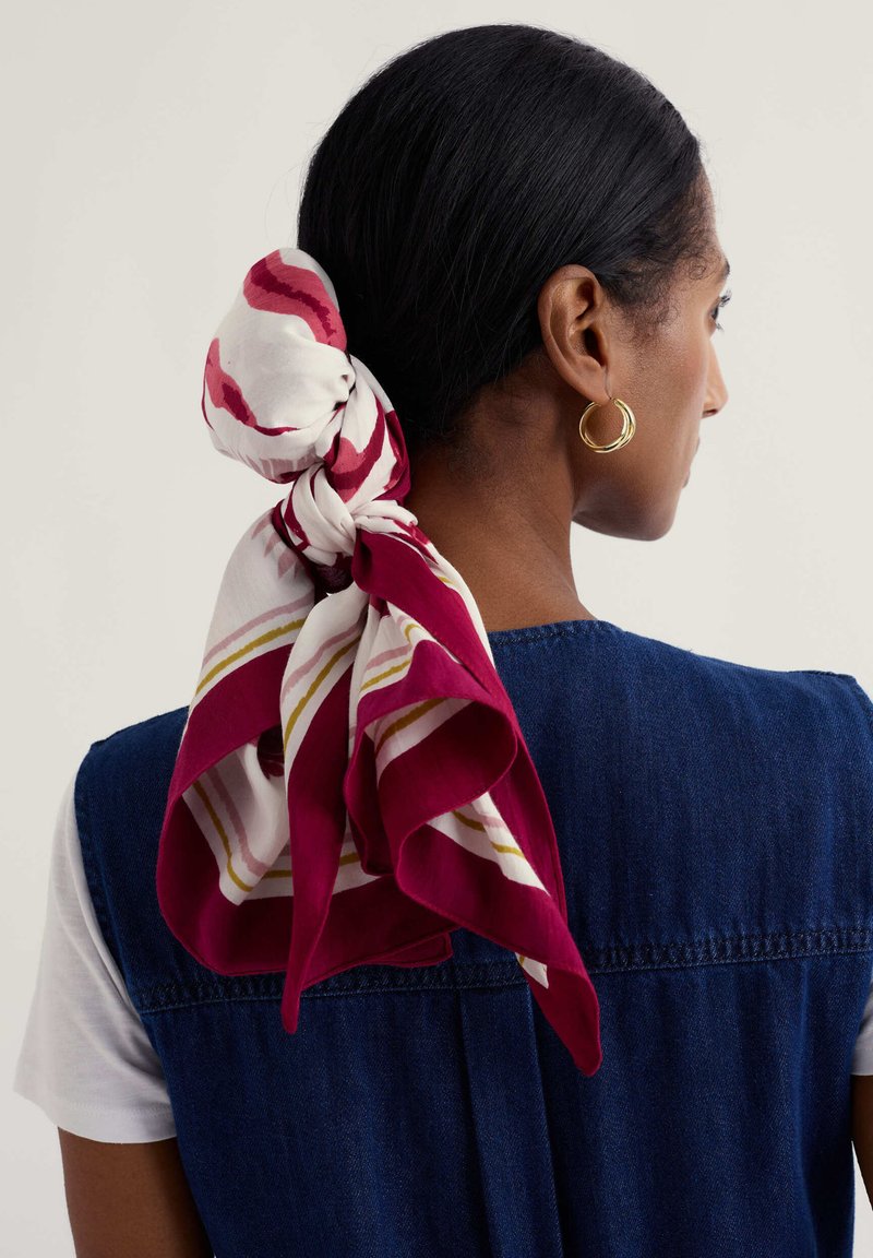 Femme aux cheveux foncés attachés avec un foulard rayé blanc et bordeaux, portant des boucles d'oreilles créoles dorées et un gilet en denim bleu par-dessus une chemise blanche.