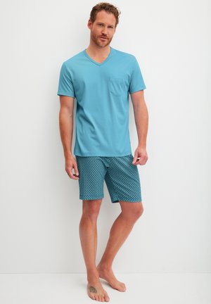 Mann steht barfuß, trägt ein blaues kurzärmliges V-Ausschnitt-Shirt und gemusterte blaue Shorts vor einem schlichten weißen Hintergrund.