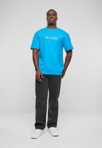 Turquoise cotton t-shirt with printed "KANI" logo, paired with black denim pants and white sneakers. Simple, short sleeves, regular fit.