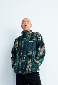 Fleecejacka med ett grönt och beigt camouflage-mönster, dragkedja fram, hög krage och en svart bröstficka med logotypdetalj.