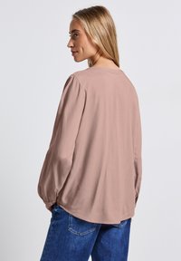 Blush-pink Bluse mit langen Ärmeln, gepufften Schultern und rundem Halsausschnitt. Weicher, glatter Stoff mit abgerundetem Saum und lockerer Passform. Jeans sind darunter sichtbar.