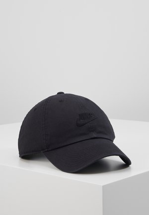 Czarna czapka bejsbolowa Nike z haftowanym logo z przodu, spoczywająca na białym sześcianie na jednolitym jasnoszarym tle.