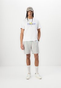 T-shirt blanche avec un logo coloré "Pepe Jeans London", short rayé vert clair et rose, baskets blanches et casquette grise.