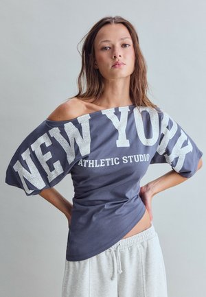 Jeune femme aux cheveux bruns lâchés porte un haut bleu foncé épaules dénudées avec le texte "New York Athletic Studio" et un pantalon de jogging gris clair.