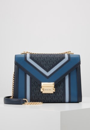 Blauwe en zwarte Michael Kors schoudertas met goudkleurige kettingriem, geometrisch patroon en rechthoekige gouden sluiting op een wit oppervlak.