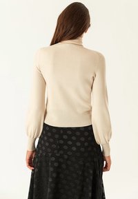 Hellbeige enganliegendes Rollkragensweatshirt mit langen, leicht puffigen Ärmeln. Weicher Stoff, gerippter Saum, kombiniert mit einem schwarzen gepunkteten Rock.