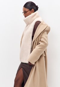 Beige vlněný kabát přes hnědý pletený top, s bílým kožešinovým límcem a hnědou koženou sukní s rozparkem na stehně. Brýle a zlaté náušnice dotvářejí celkový vzhled.