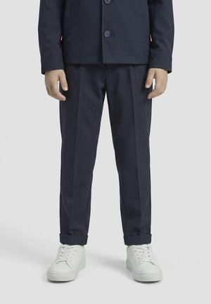 Osoba stoji u mornarskoplavim krojenim hlačama s manžetama, odgovarajućem saku i bijelim tenisicama na svijetlosivoj pozadini.