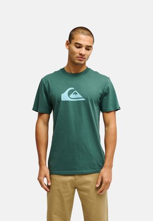 Jeune homme aux cheveux courts portant un t-shirt vert avec un logo vague bleu clair et un pantalon beige, debout devant un fond uni.