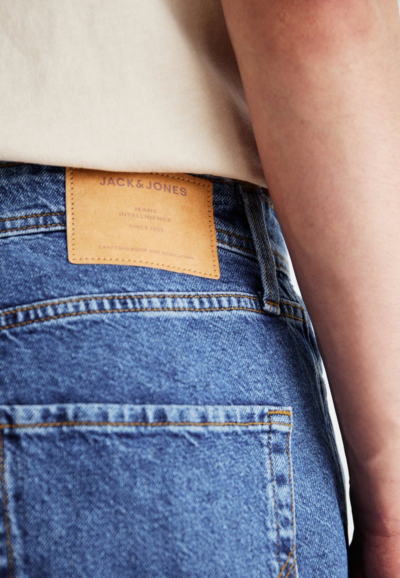 Jeans in denim blu con un'etichetta in pelle con la scritta "JACK & JONES" sulla vita. Presentano cuciture visibili e tasche posteriori.