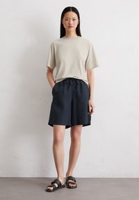 Beige T-shirt met korte mouwen gecombineerd met donkerblauwe shorts met trekkoord en zwarte sandalen met gouden accenten. Glad stof, comfortabele pasvorm.