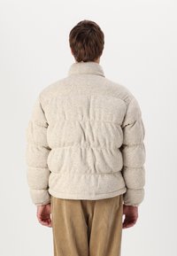 Puffjacka i ljusbeige med texturerat tyg, med hög krage och rynkade sektioner. Matchad med bruna kordflosbyxor.