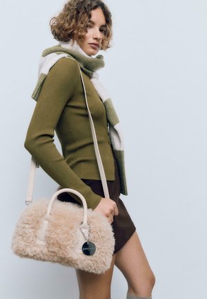 Junge Frau in einem grünen Pullover und gestreiftem Schal hält eine beige, flauschige Handtasche, trägt einen braunen Rock und steht vor einem einfarbigen Hintergrund.