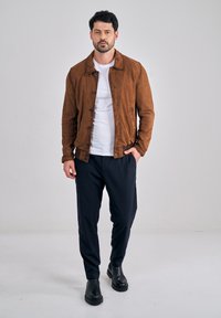 Man met donker haar die een bruine suède jas, wit T-shirt, zwarte broek en zwarte laarzen draagt, staand tegen een eenvoudige lichte achtergrond.