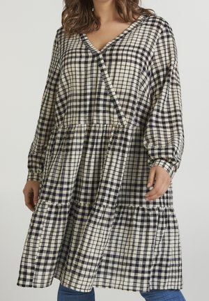 Femme portant une robe ample à manches longues à carreaux noirs et blancs avec une jupe froncée par-dessus un jean bleu.