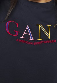 T-shirt in cotone blu navy con testo ricamato in rosso, rosa e giallo che recita "GANT AMERICAN SPORTSWEAR" sul petto.
