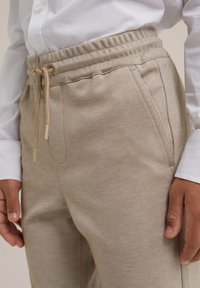 Pantalones color beige con cordón de ajuste en un tejido texturizado, con bolsillos laterales y cintura elástica, combinados con una camisa blanca.