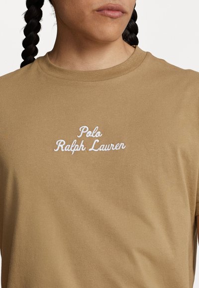 Béžové bavlnené tričko s rebrovanou textúrou, s bielym vyšitým textom "Polo Ralph Lauren" na prednej strane. Okrúhly výstrih a krátke rukávy.