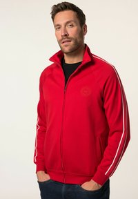 Giacca rossa con zip e strisce bianche ai lati, polsini e colletto a costine. Presenta un logo sul petto e due tasche frontali. Tessuto liscio.