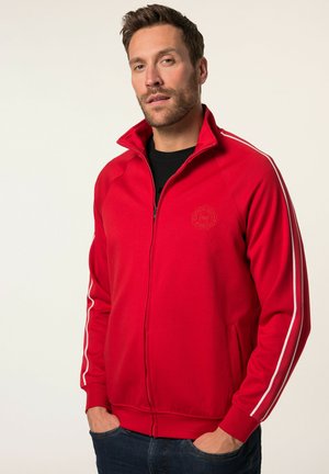 Chaqueta roja con cierre de cremallera y rayas blancas en los laterales, puños y cuello acanalados. Presenta un logo en el pecho y dos bolsillos frontales. Textura de tela suave.