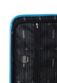 Intérieur noir d'une valise avec une fermeture éclair bleue, ornée de motifs d'avions et d'une étiquette avec des instructions d'entretien. Tissu texturé et poche en maille.